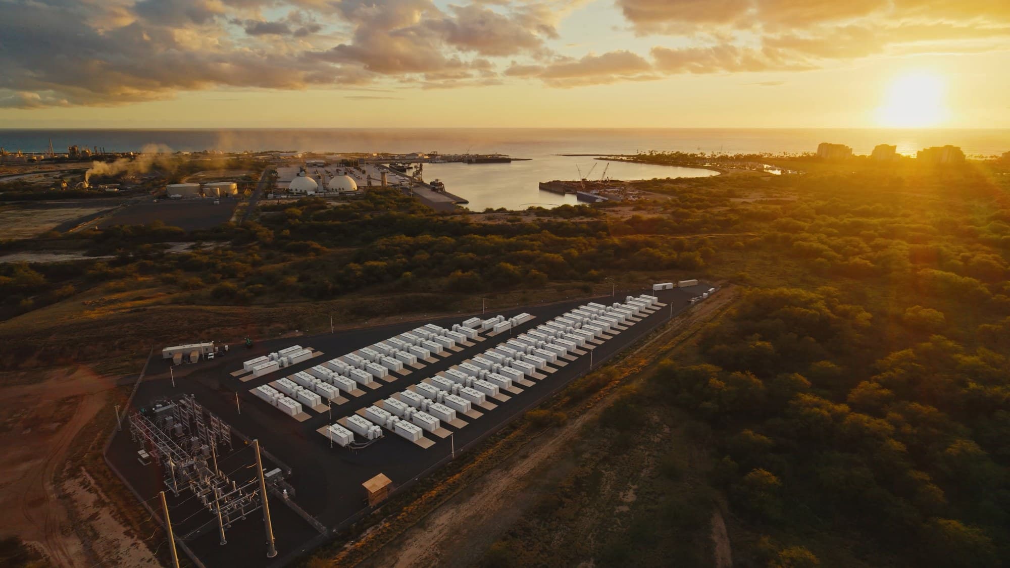 Rows of white Tesla Megapack battery units at a grid storage site beside a substation at sunset