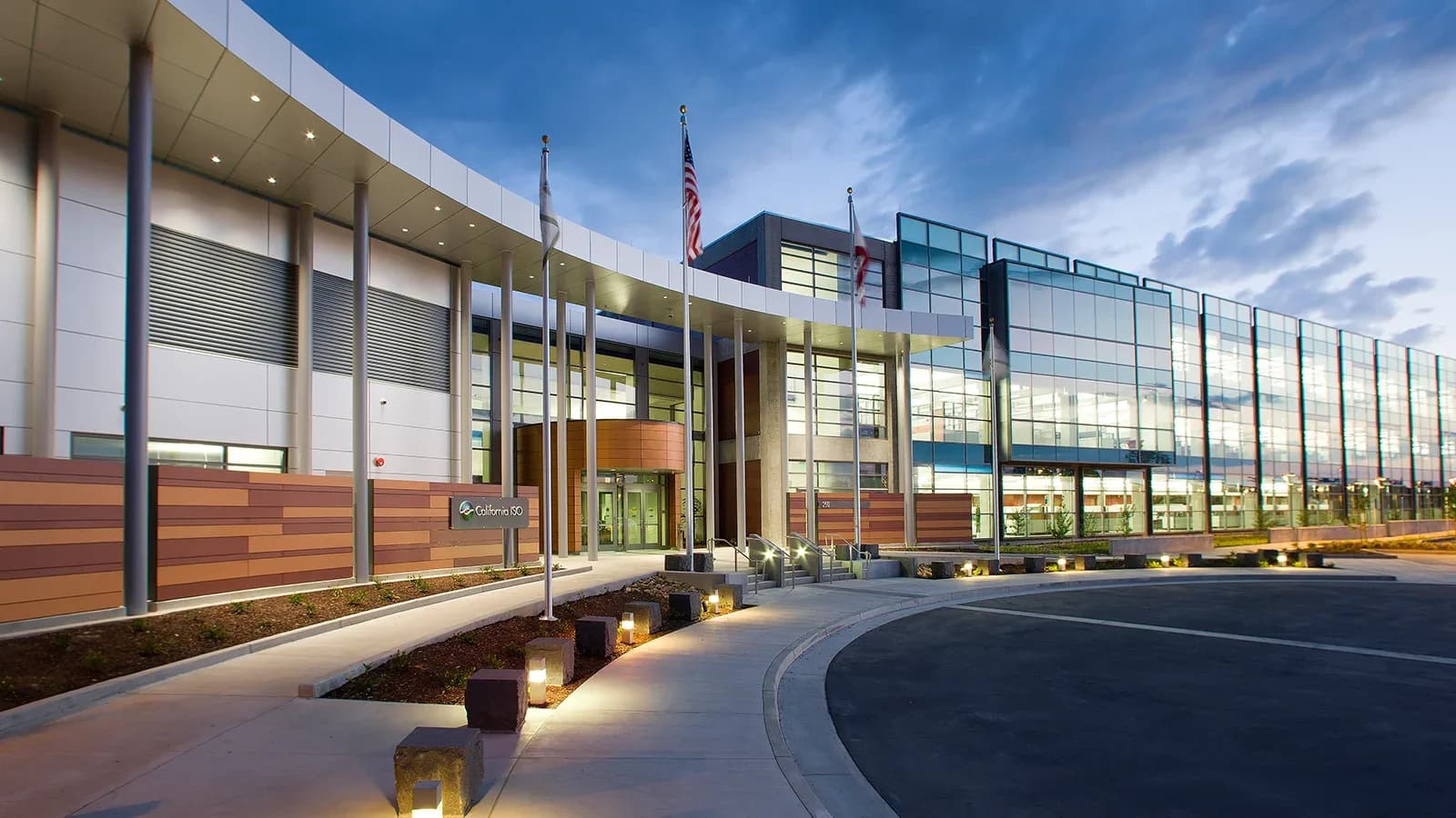 Entrance and glass-fronted office building of the California ISO headquarters at dusk, with the California ISO sign beside the walkway