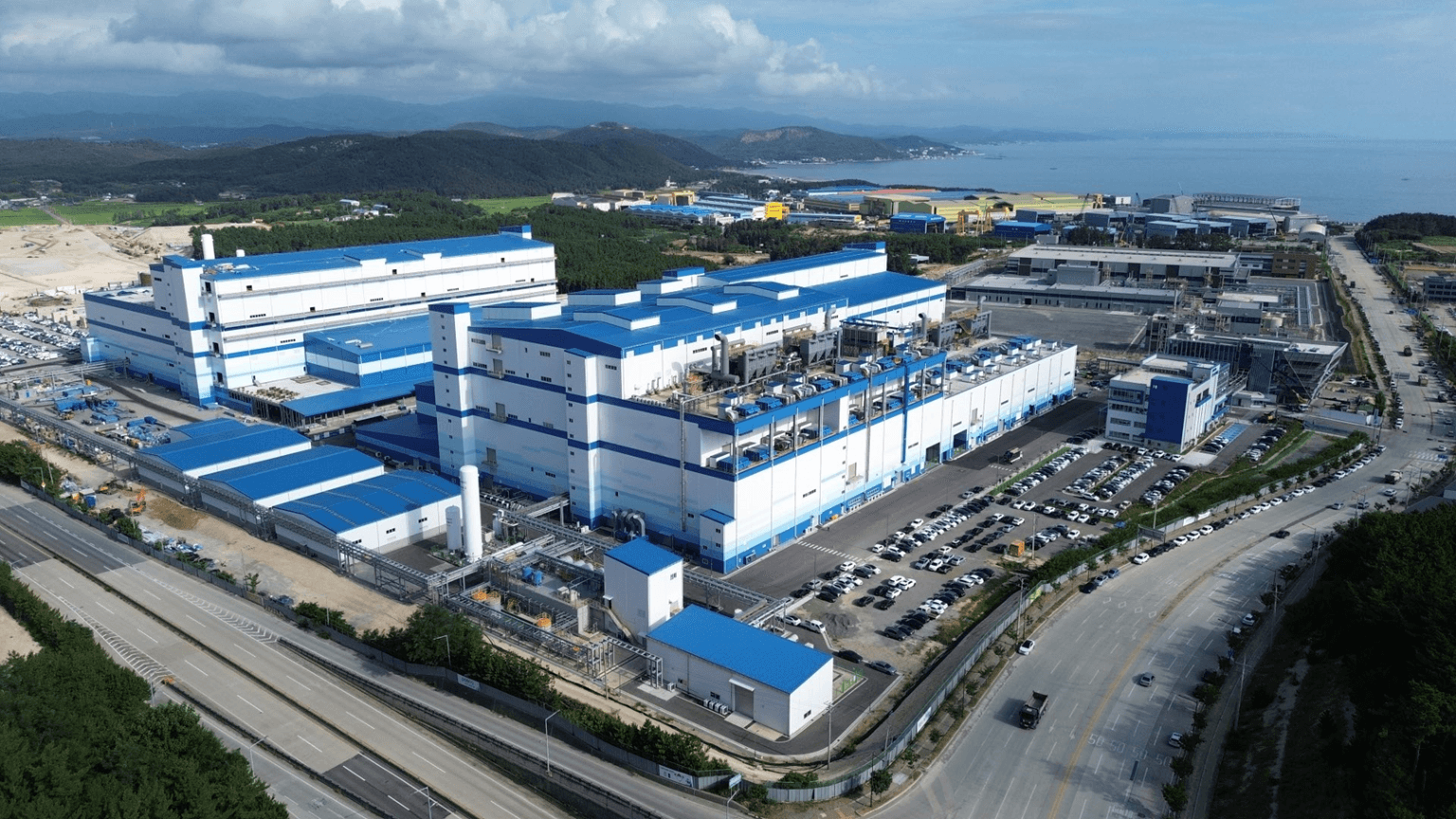 Aerial view of a POSCO Future M cathode materials plant with blue-and-white production buildings beside a coastal road in South Korea