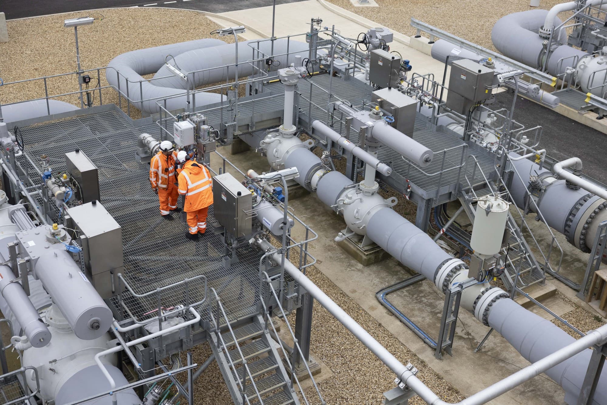 Grey insulated gas pipework, valves and walkways at a National Gas Transmission site, with two workers in orange high-visibility jackets