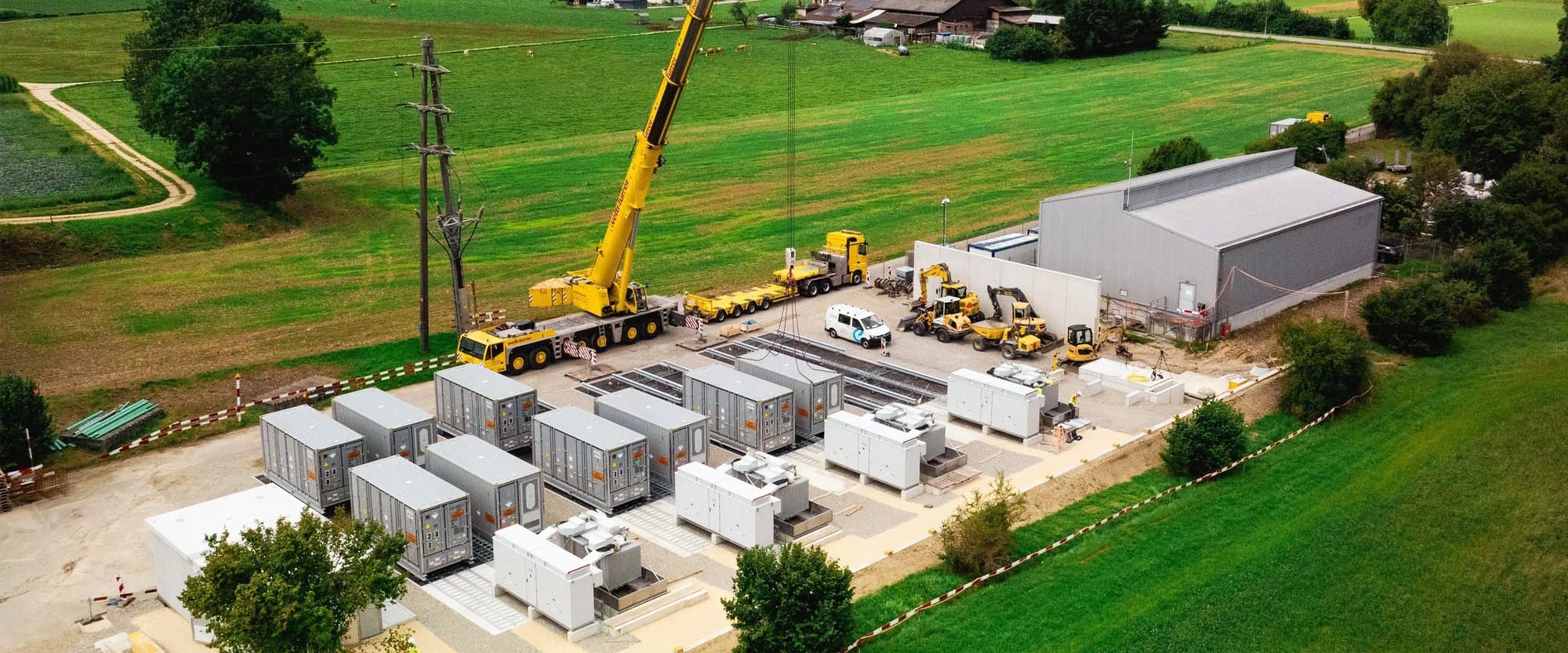 Aerial view of grey battery storage containers, inverters and a crane at an Intilion storage site beside green fields