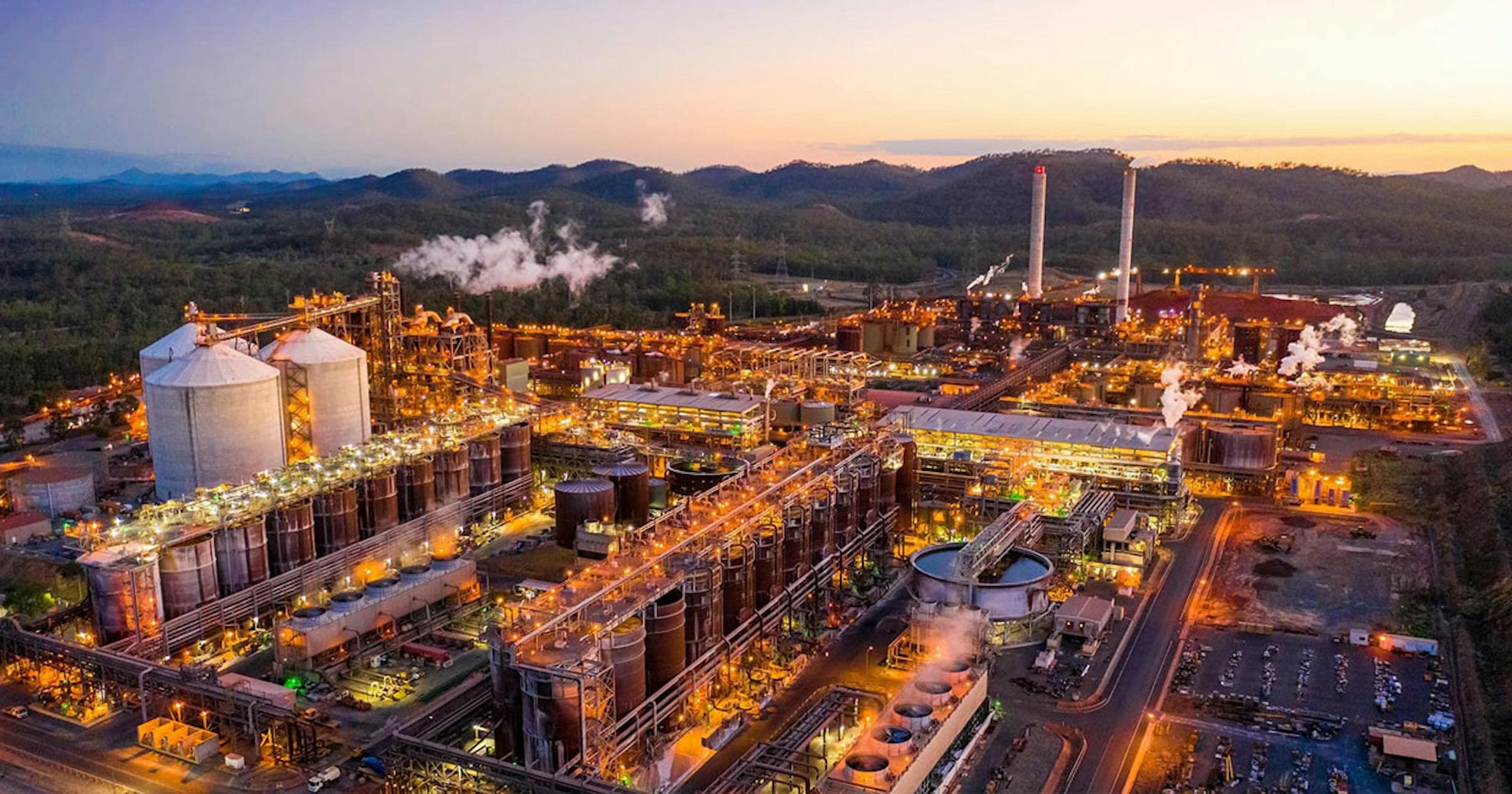 Aerial view at dusk of an alumina refinery with lit process tanks, silos and steam plumes, from Rondo Energy's Rio Tinto release