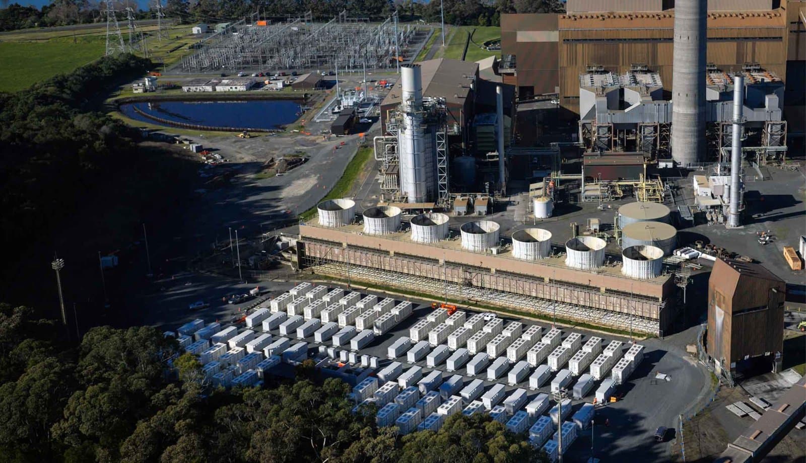 Aerial view of rows of white battery containers beside the cooling towers of Huntly Power Station in New Zealand.