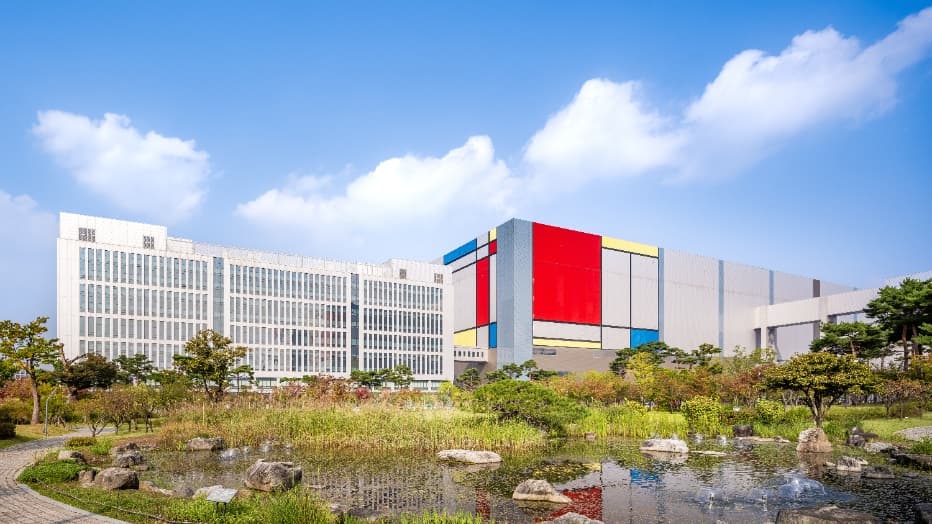 Samsung's Pyeongtaek semiconductor campus with a colorful fab building behind a garden pond on a clear day.