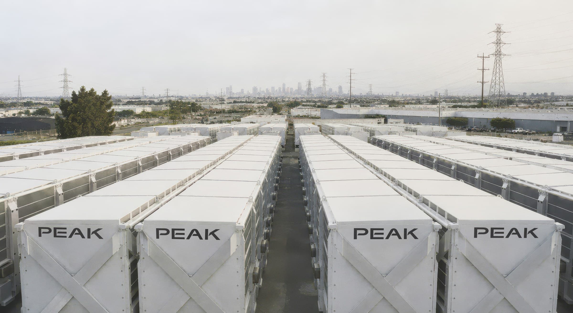 Rows of white Peak-branded sodium-ion energy storage enclosures lined up outdoors with a city skyline in the distance