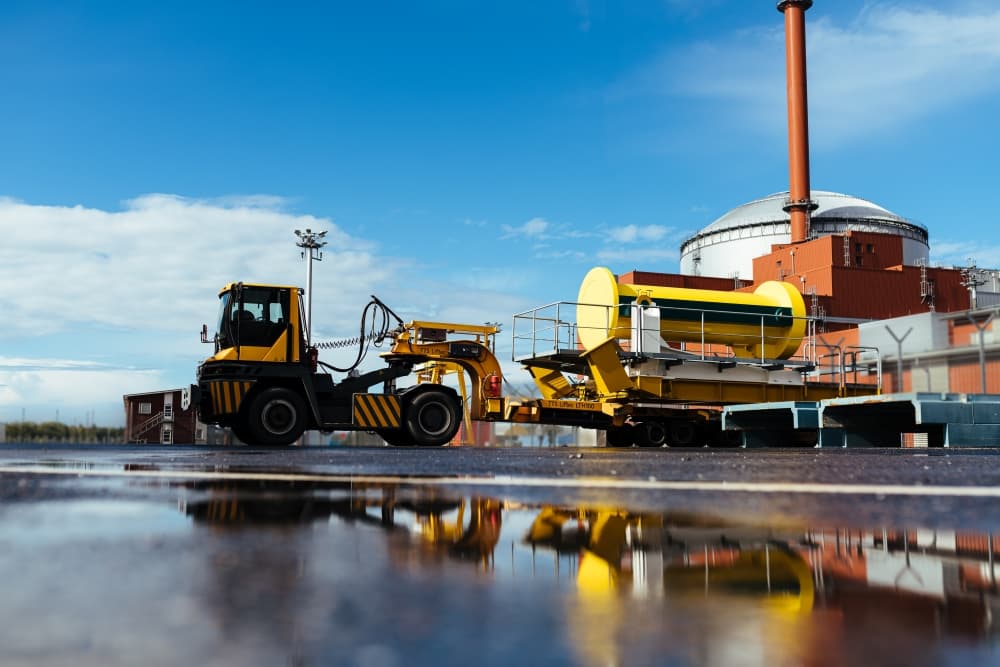 Yellow spent-fuel transfer cask on a trailer at Posiva's Olkiluoto site in Eurajoki, Finland, with a reactor building behind